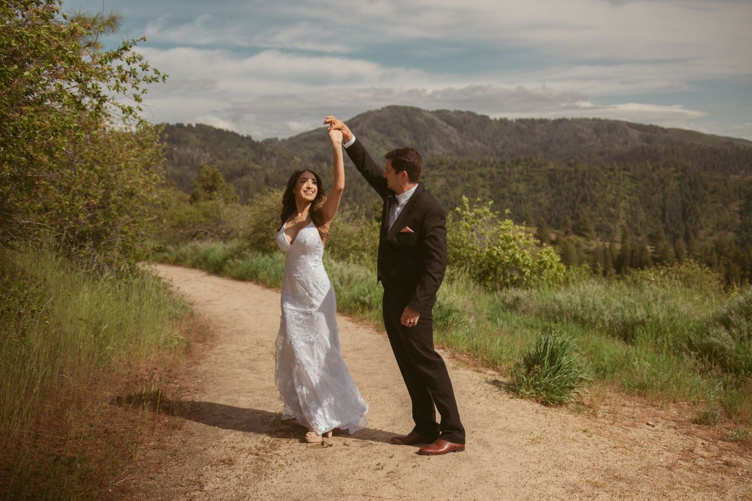 A couple shares a dance moment on a mountain hiking trail during a sunny day.