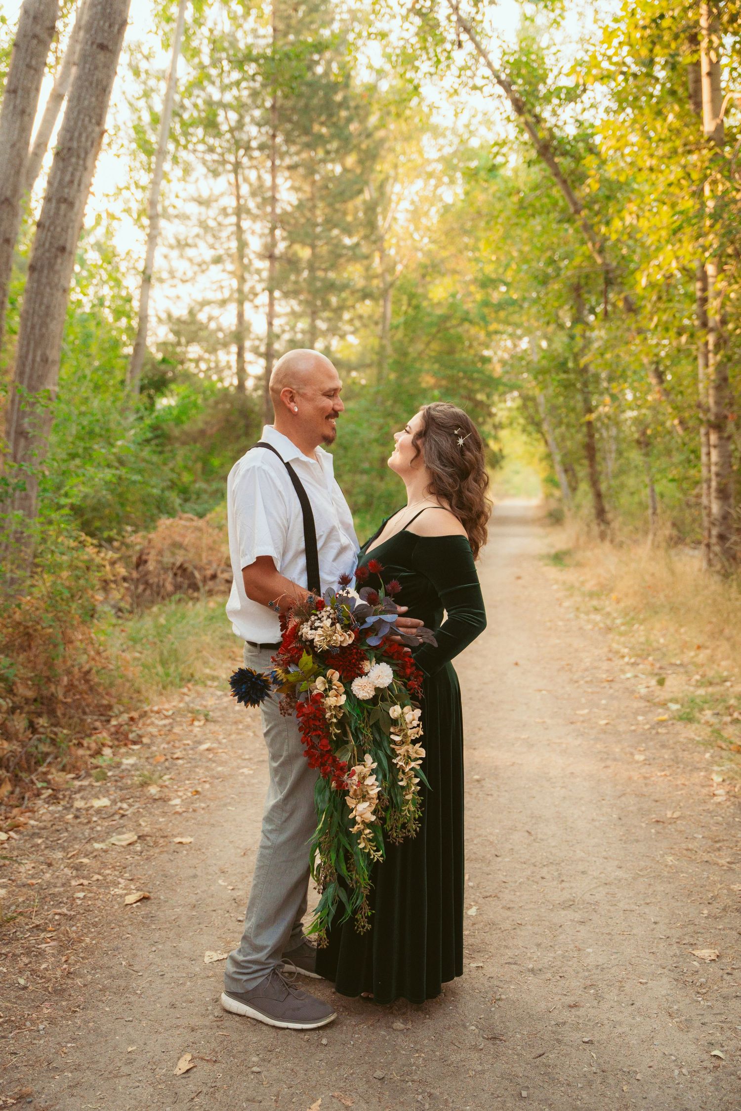 A romantic couple poses on a wooded path during golden hour with autumn foliage and a beautiful floral bouquet.