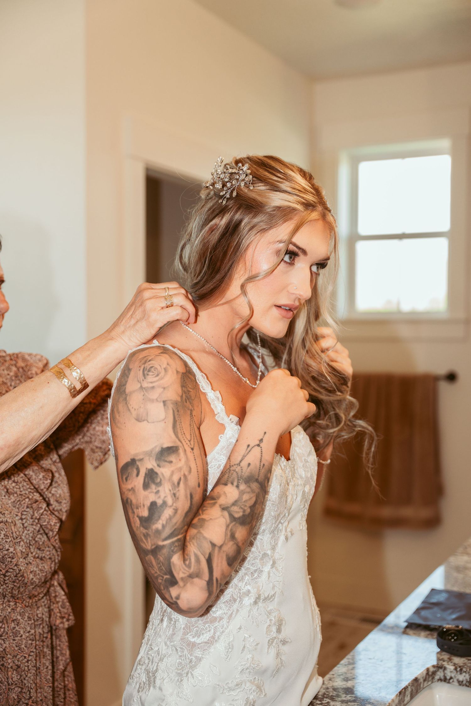A bride in a white lace dress with visible arm tattoos gets ready on her wedding day.