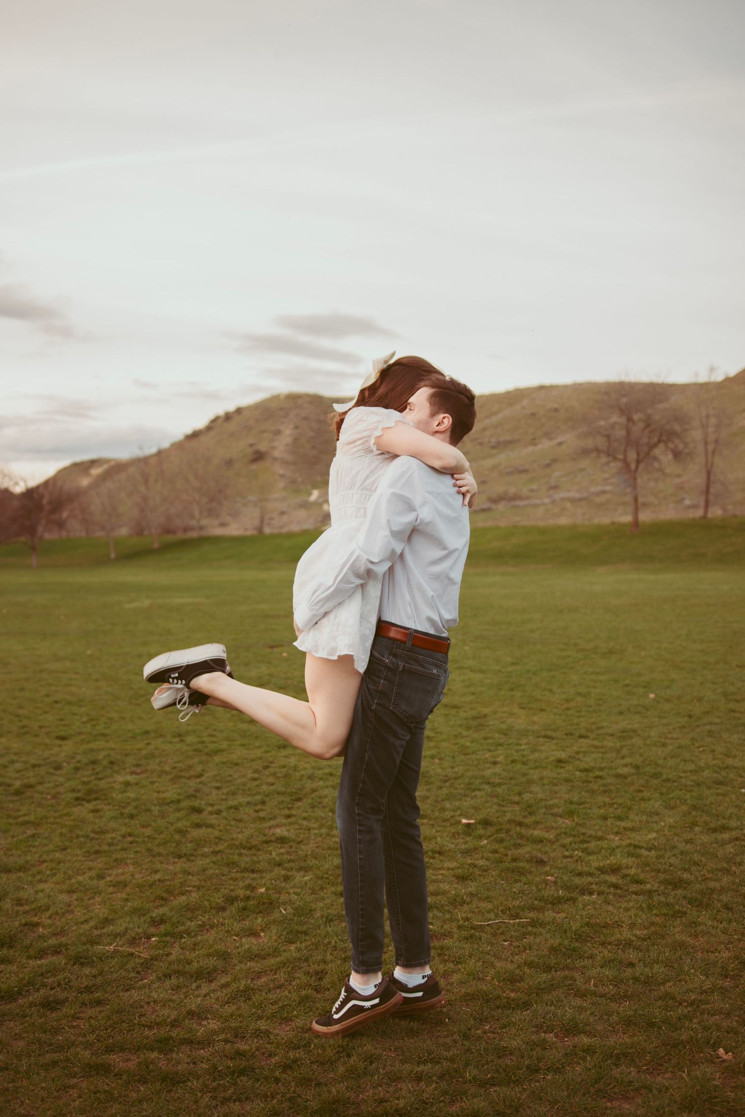 Joyful couple embraces in grassy field with rolling hills in background at sunset.