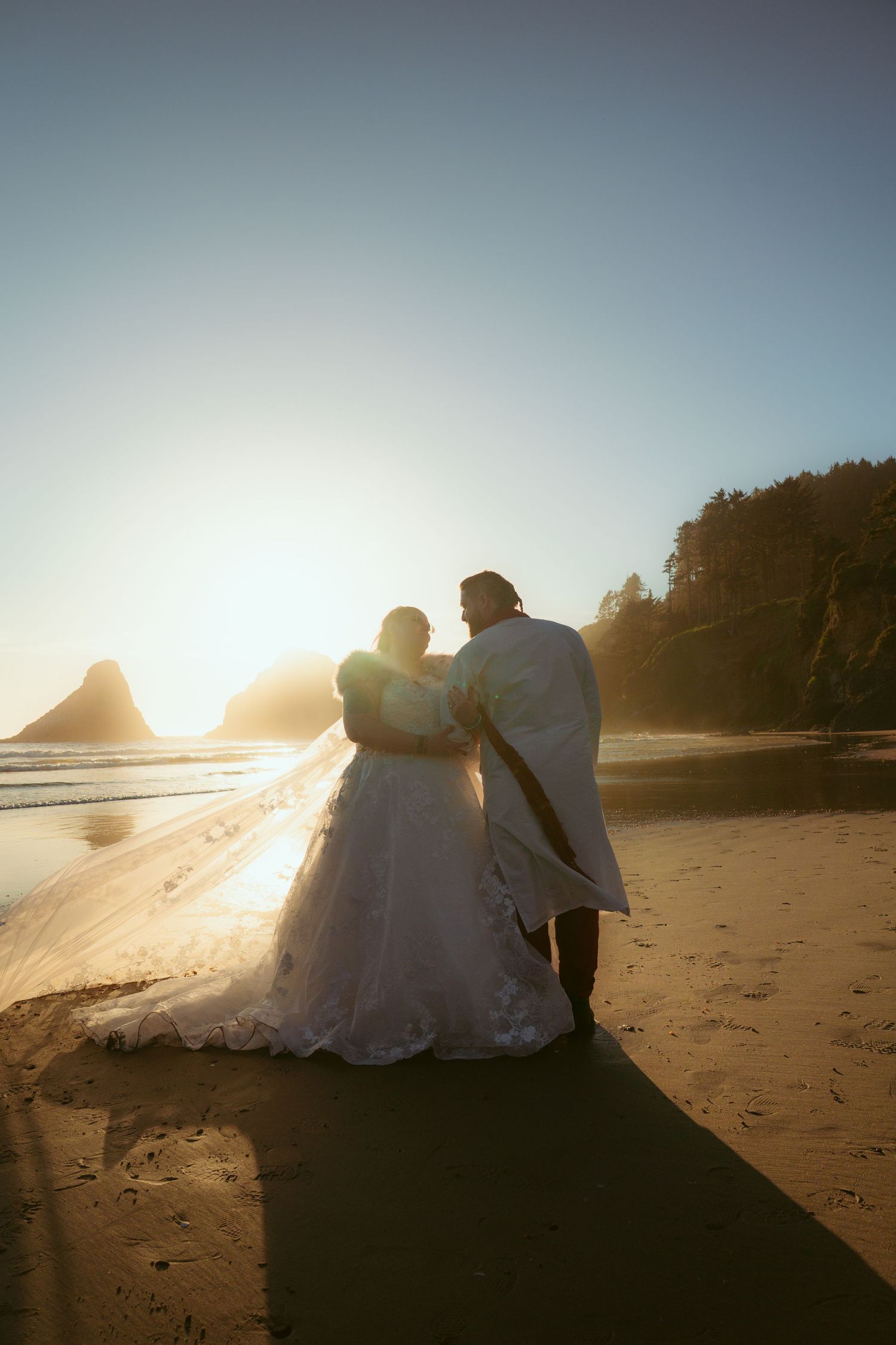 A couple in wedding clothes embraces on a sandy beach during sunset with coastal cliffs in the distance.