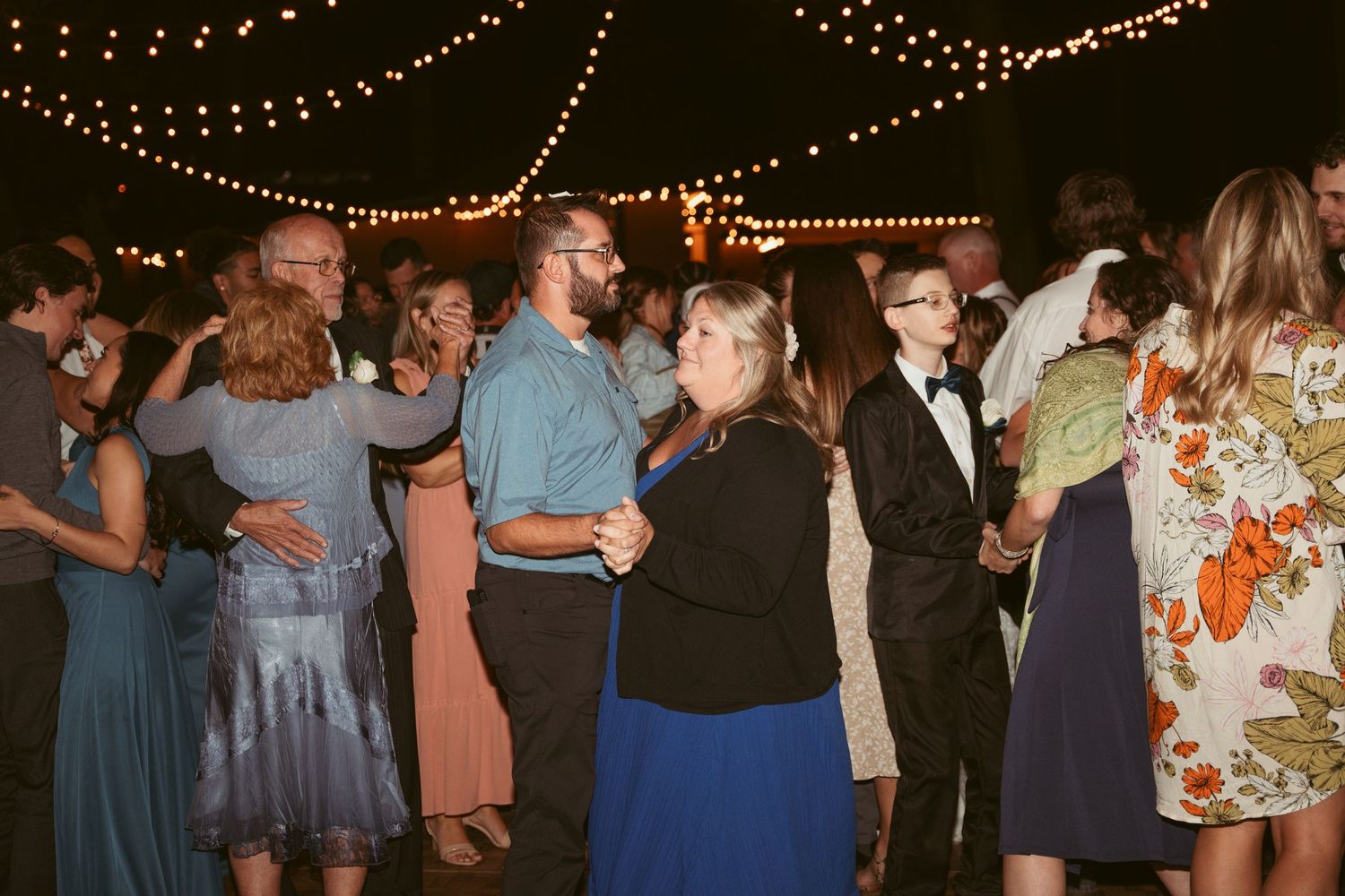 Crowd of party guests dancing and mingling under warm string lights at night.
