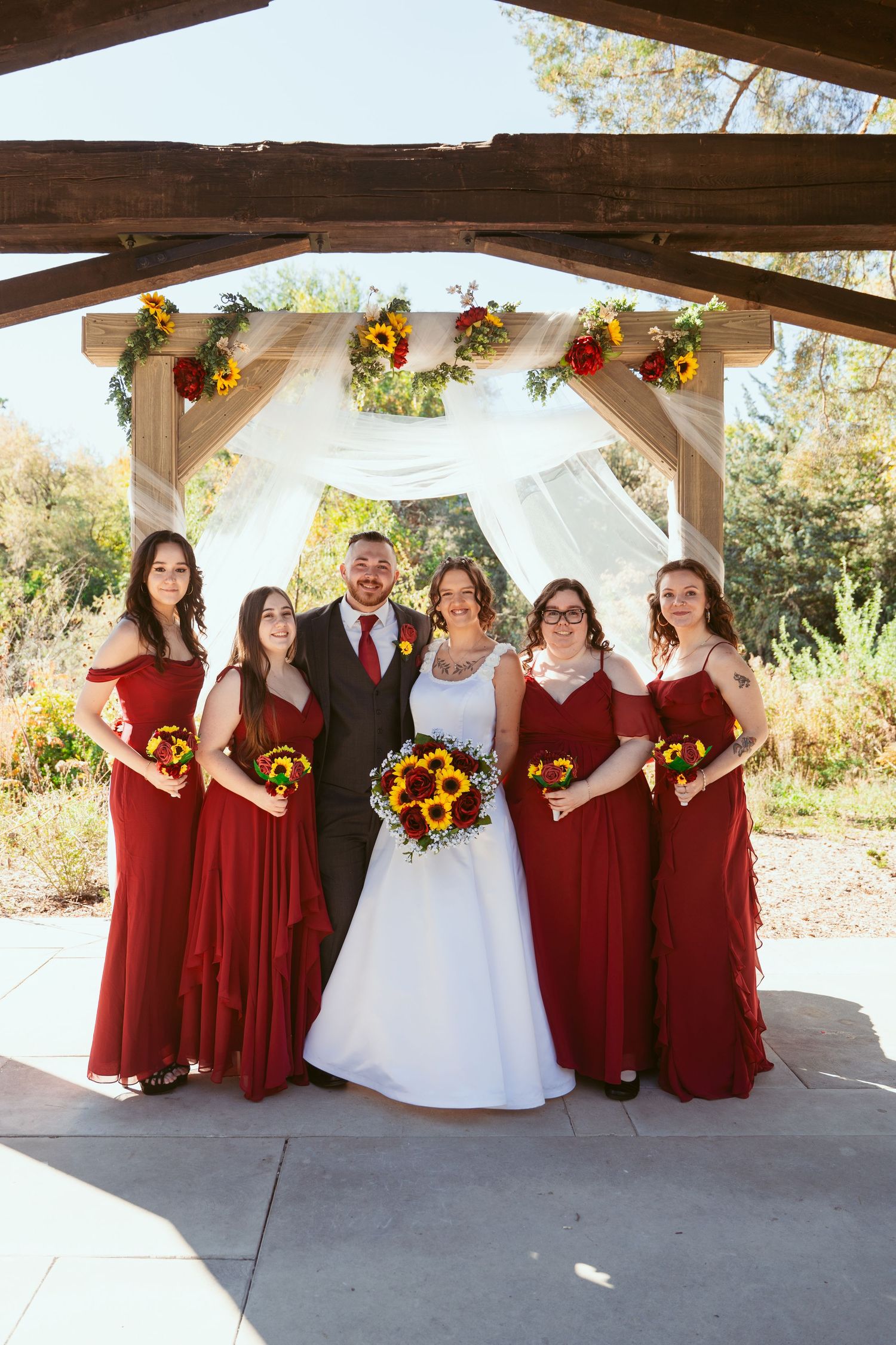 Wedding party in burgundy and white poses with sunflower bouquets under wooden pavilion.