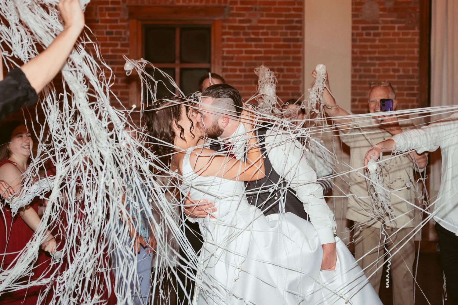 Group of wedding guests throwing white streamers during reception party in brick-walled venue.