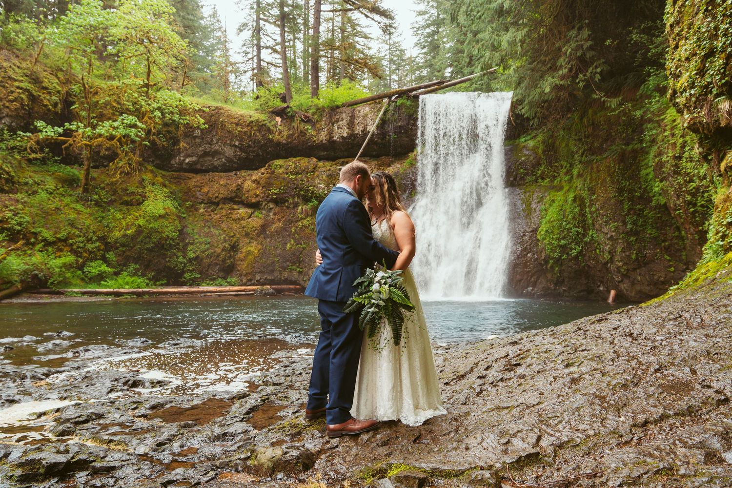 Romantic wedding photoshoot along a rocky riverbank with a majestic waterfall in the background.