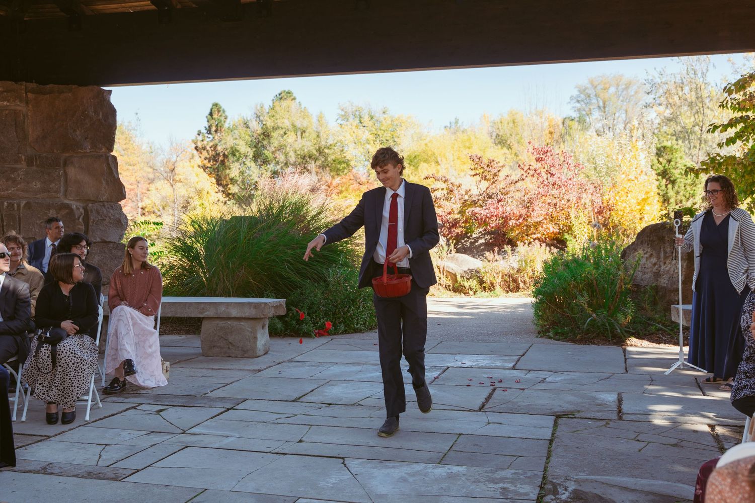 Wedding ceremony participant walks down aisle carrying red basket outdoors.