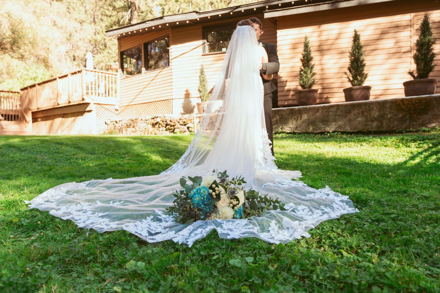 Wedding dress with long veil spread out on grass with bouquet in front of rustic wooden building.