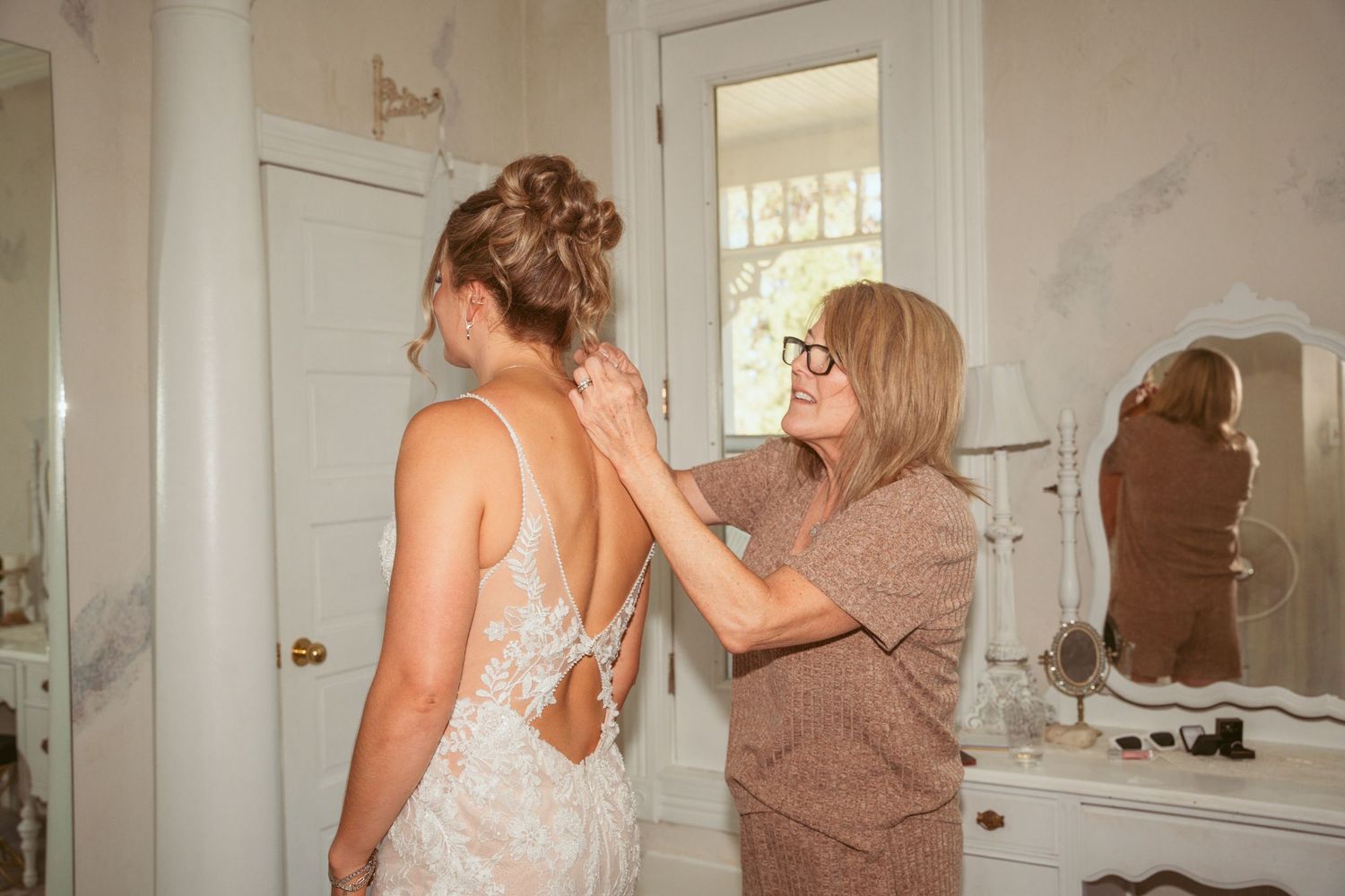 Helper assisting with the back of a delicate lace wedding gown during final preparations.