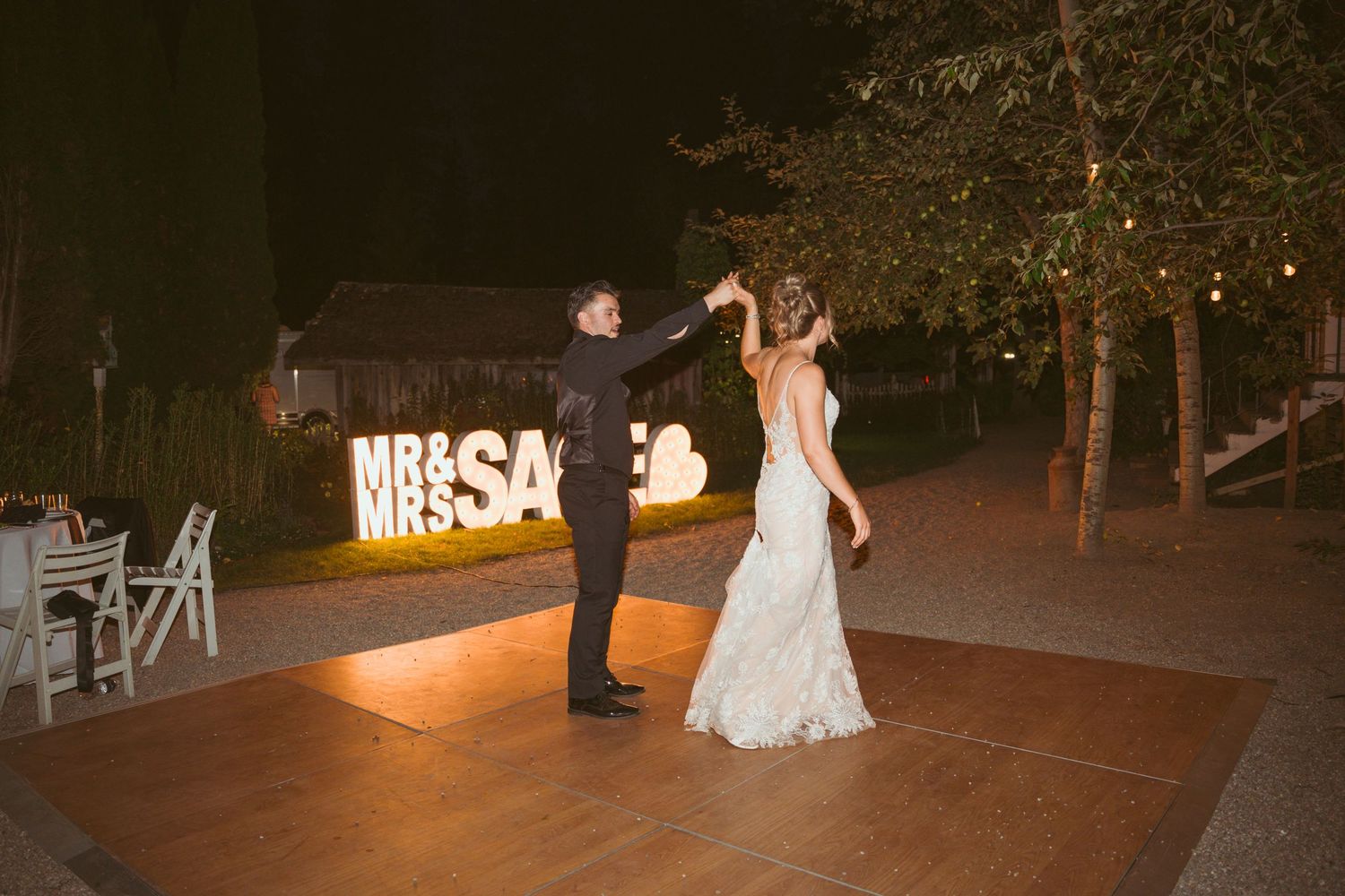 Couple dancing together in front of illuminated 'MR & SAGE' sign at outdoor reception.