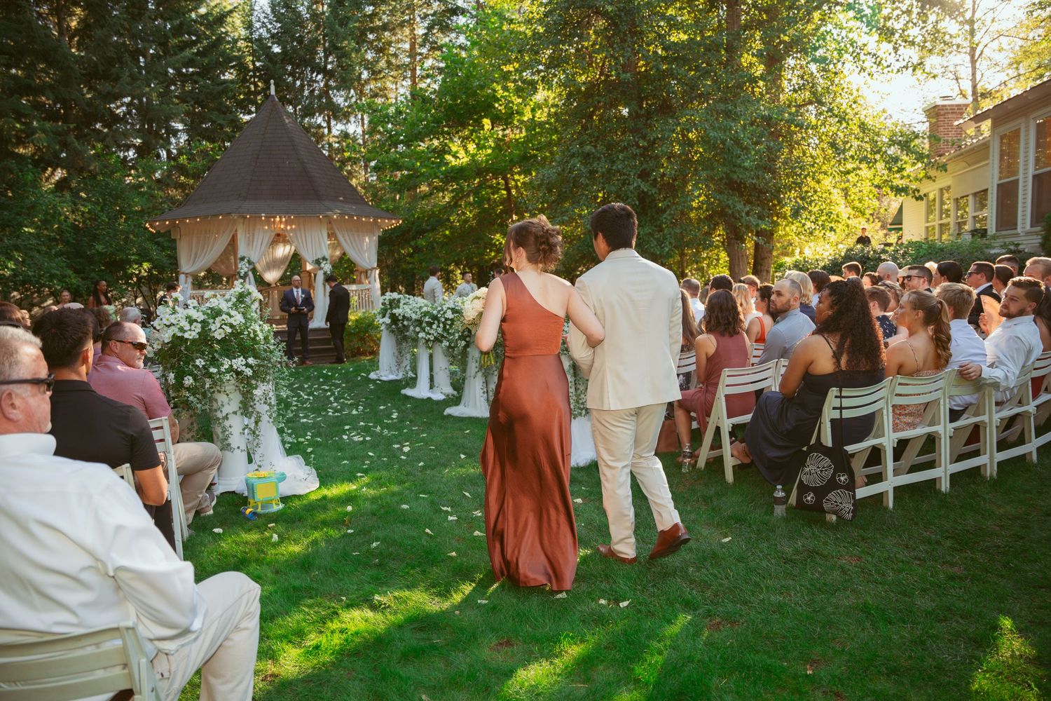 Garden wedding venue with white gazebo and guests seated on lawn during golden hour.