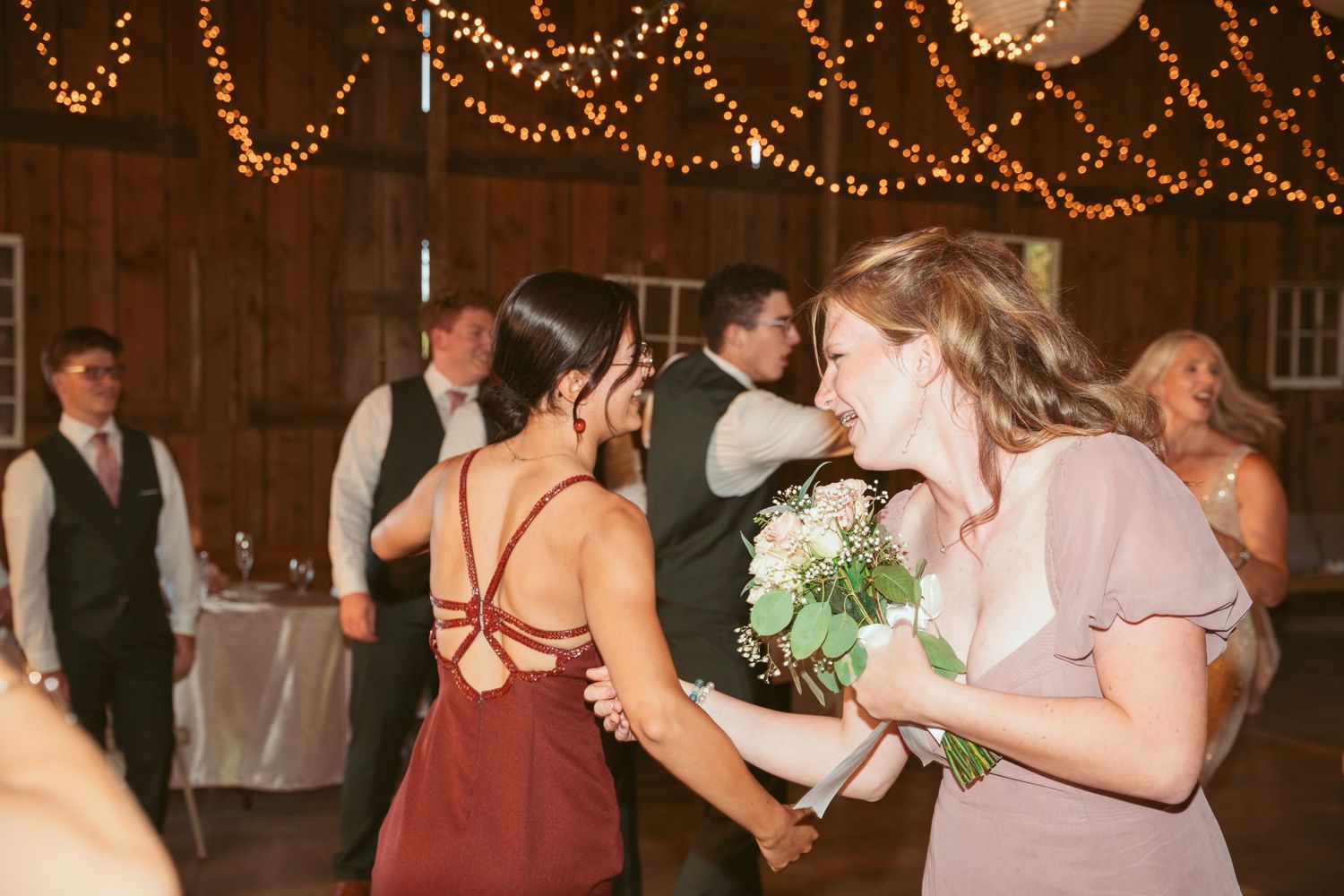 Wedding guests dance and celebrate under string lights at a rustic barn reception.