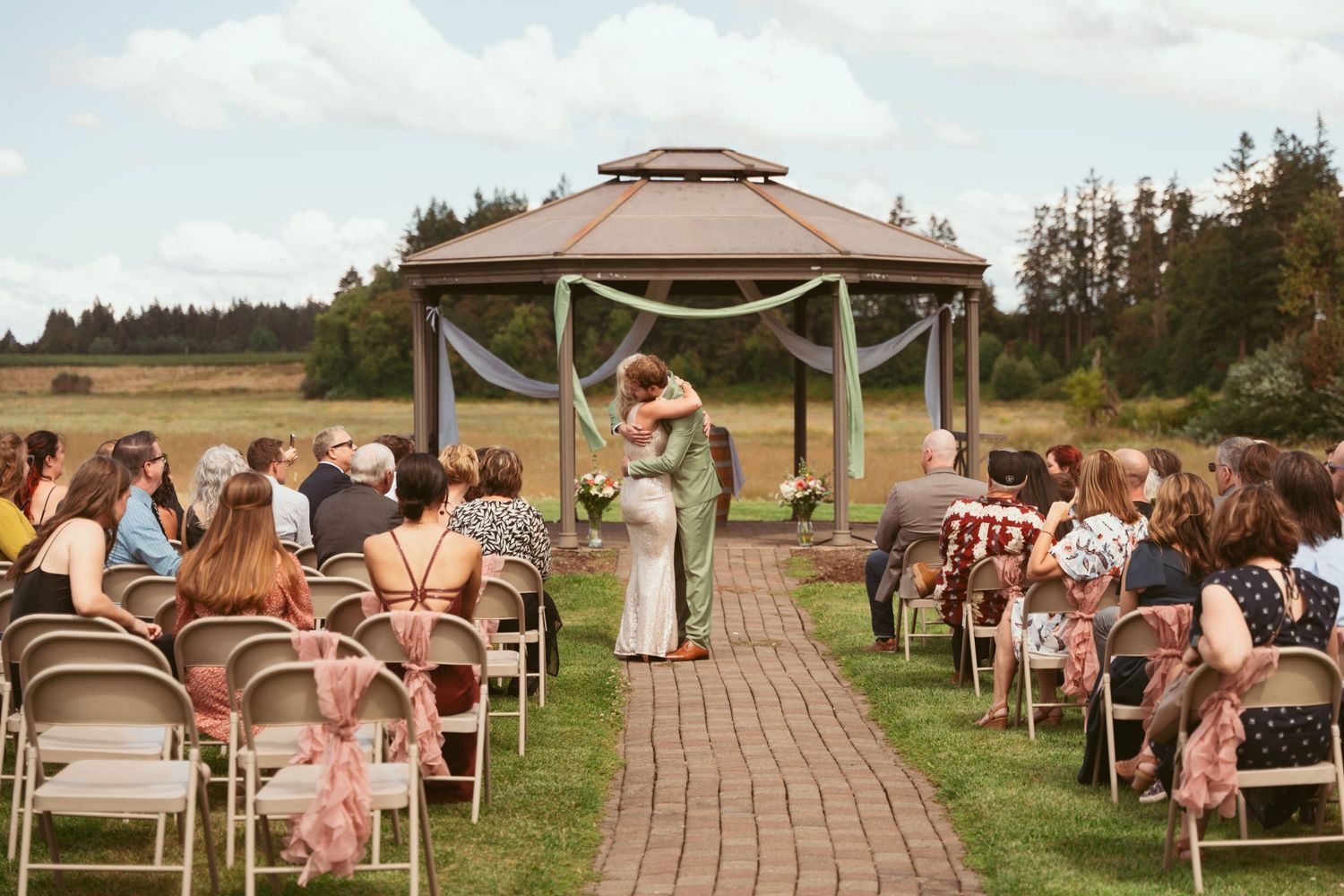 Outdoor wedding ceremony setup with brown gazebo and rows of chairs on brick pathway surrounded by green lawn.