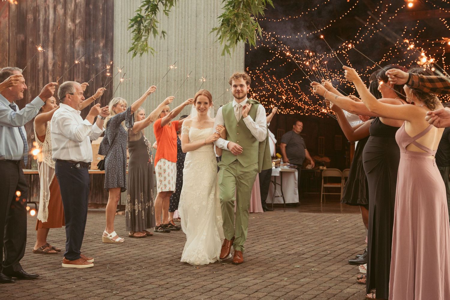 Wedding party creates a sparkler exit tunnel on brick patio with rustic barn backdrop.