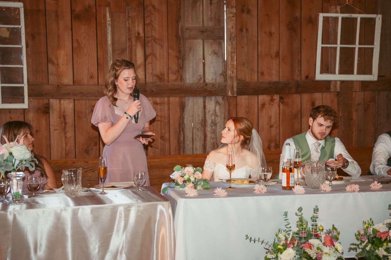 Long head table setup with floral decorations and white linens in rustic barn setting.