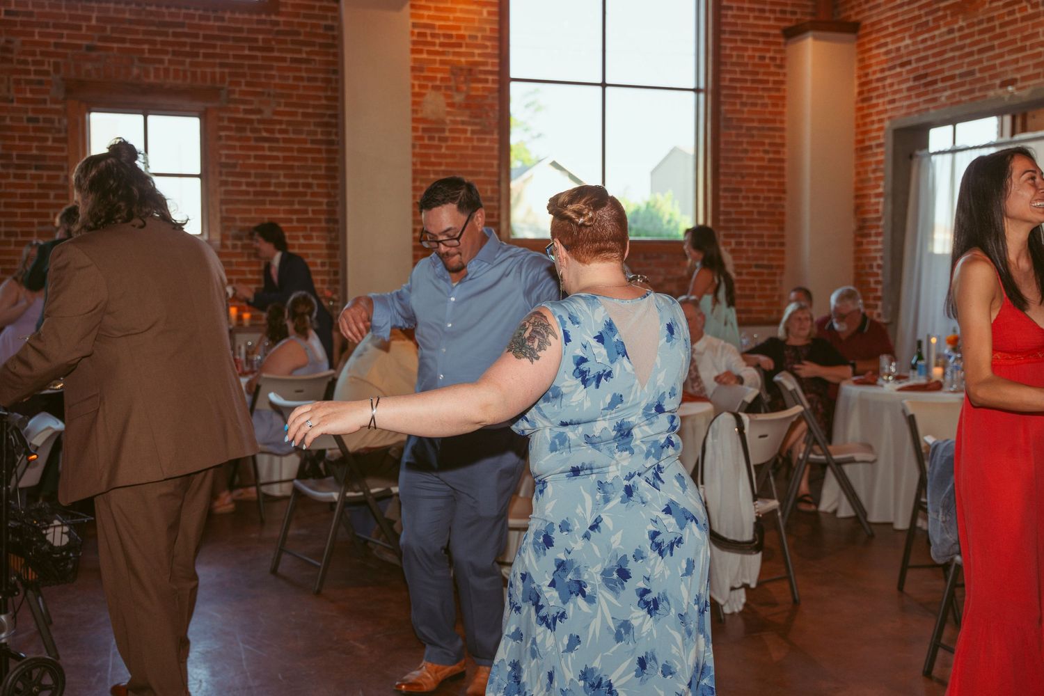 Guests dance together during a wedding reception in a sunlit brick venue with folding chairs.