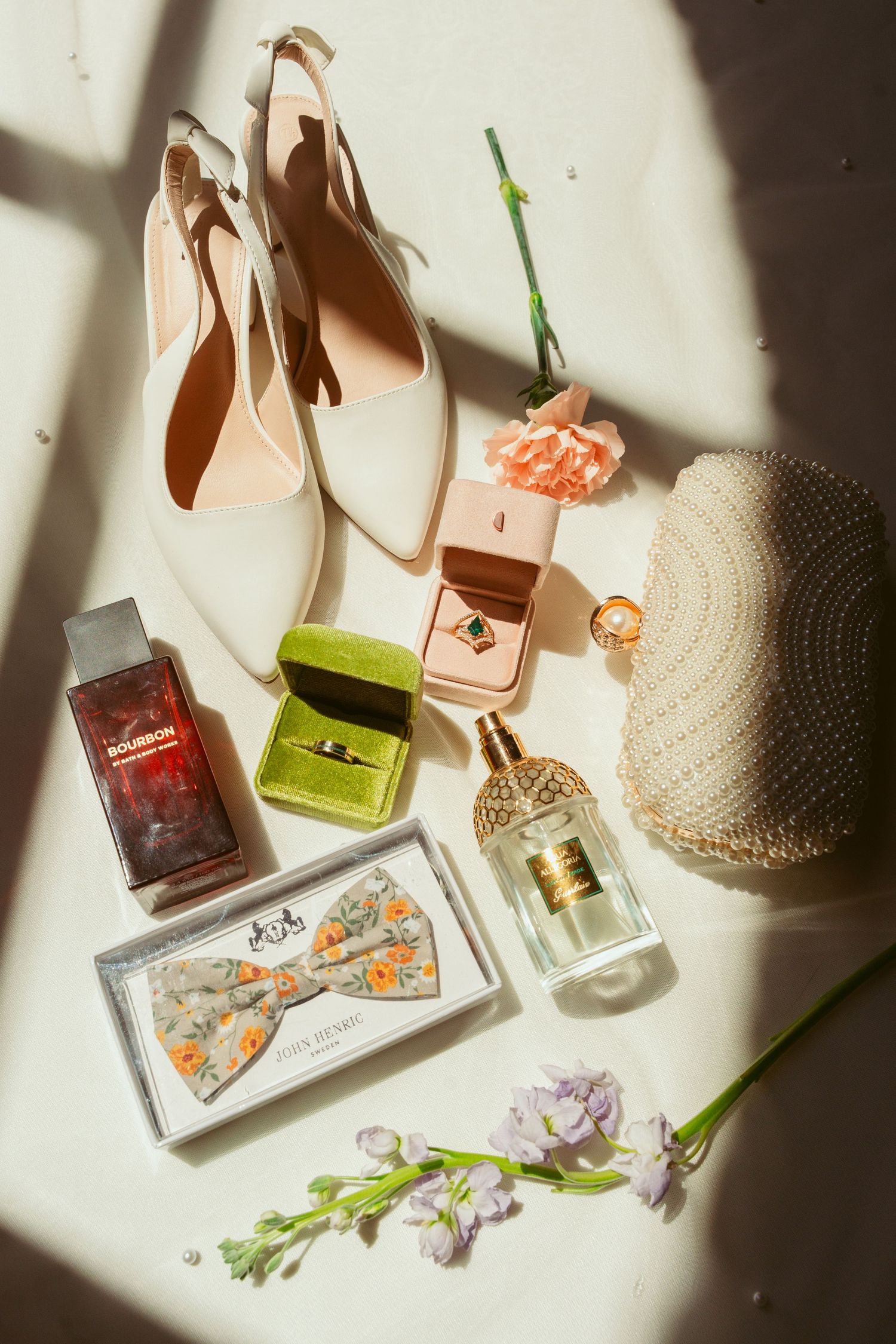 Flatlay of wedding accessories including shoes, perfume, bow tie and straw hat in sunlight.