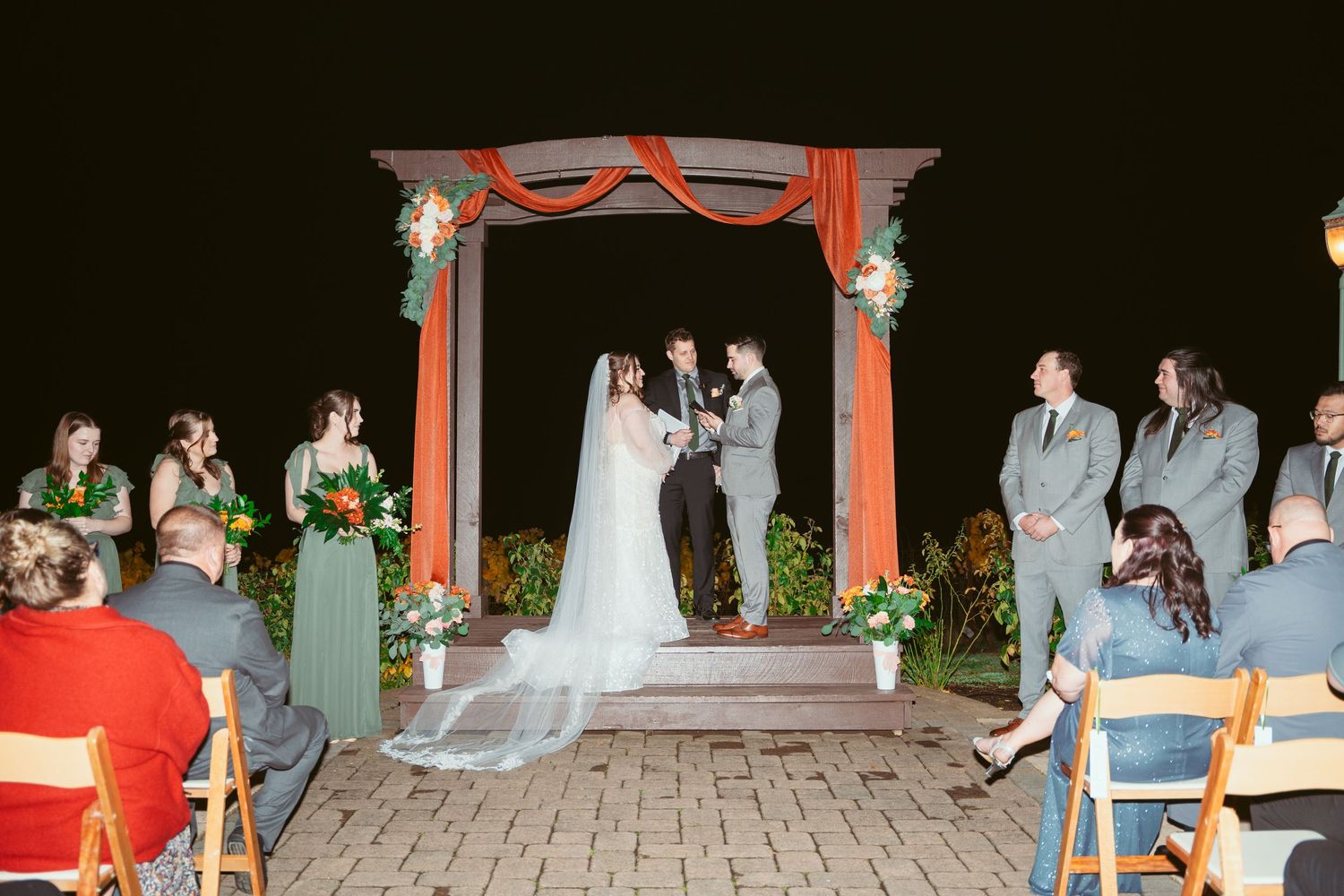 Wedding ceremony under a red-draped pavilion with guests seated on wooden chairs at dusk.