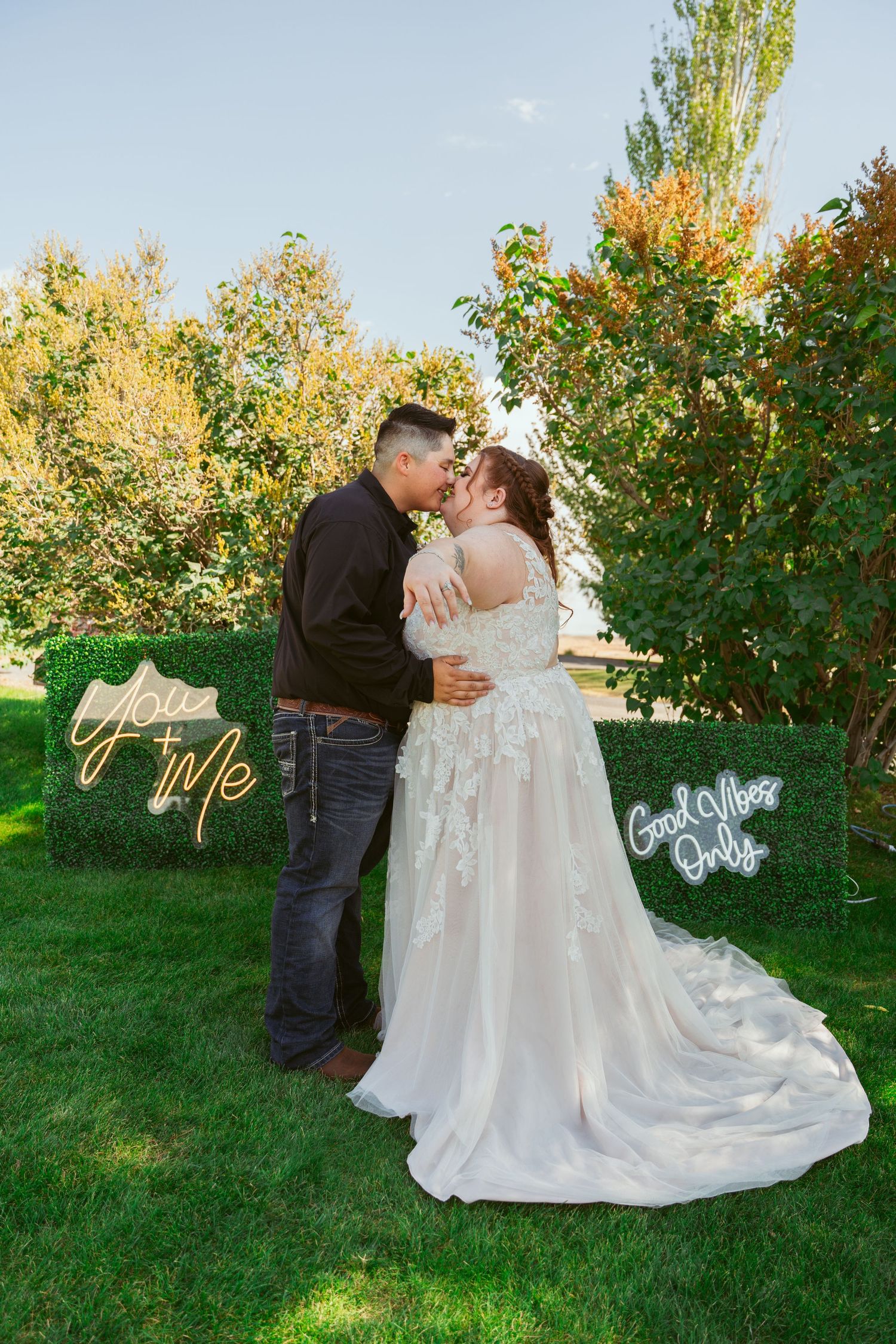 Wedding couple poses together on grass with decorative signage and trees in an outdoor garden setting.
