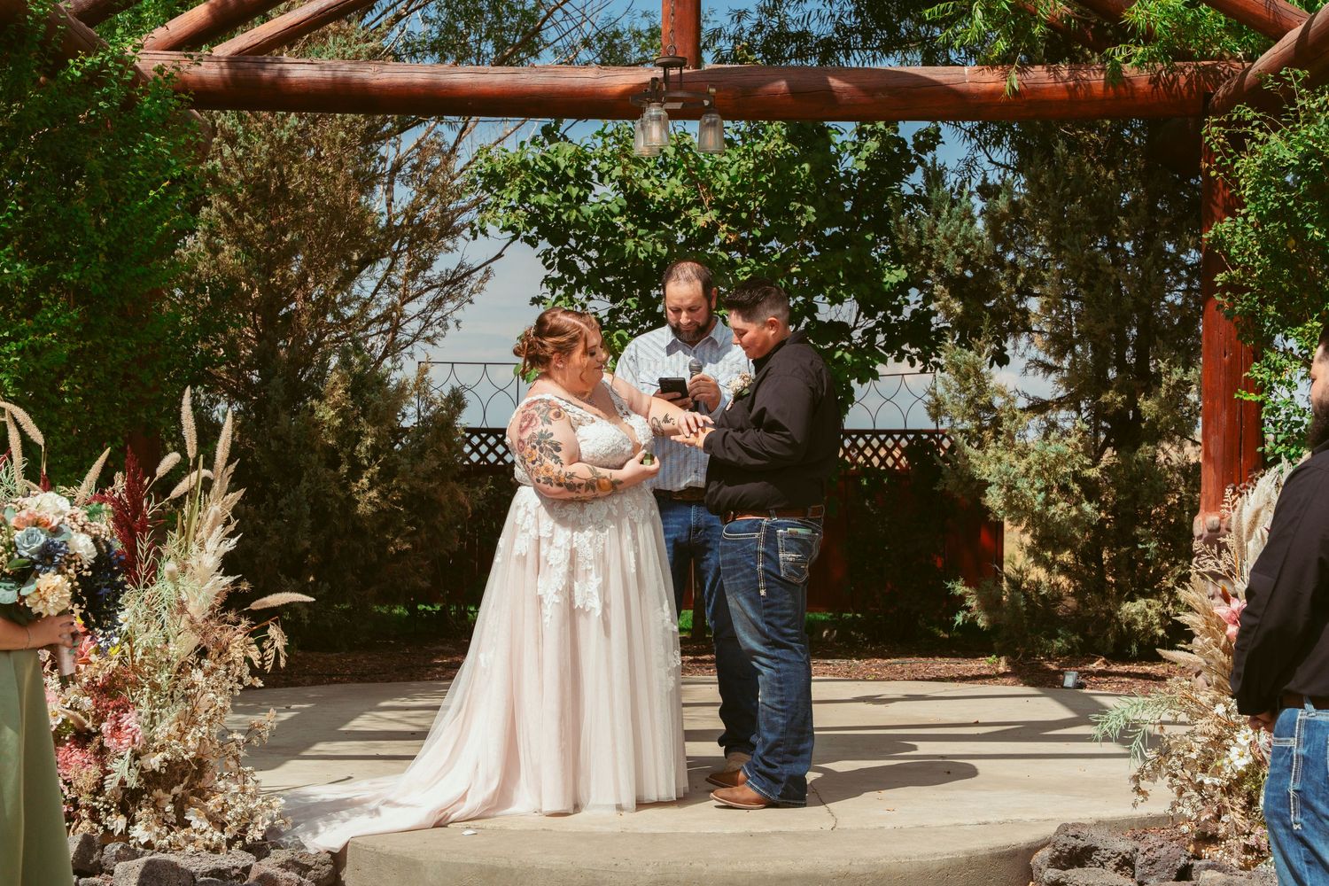 An outdoor wedding ceremony unfolds beneath a wooden pergola structure with natural landscaping and floral decorations.