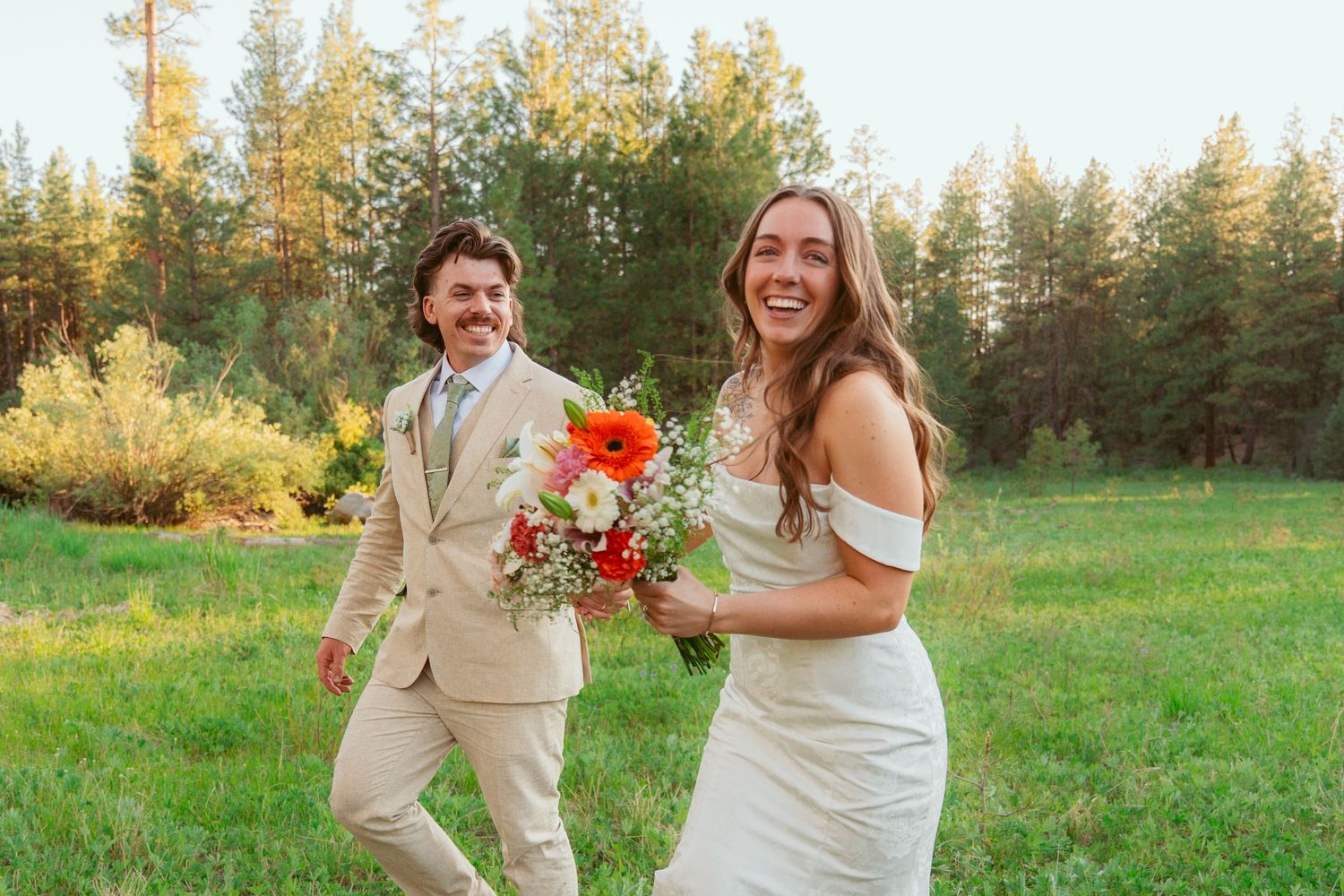 Wedding couple shares laughs while holding bouquets in a sunny meadow.