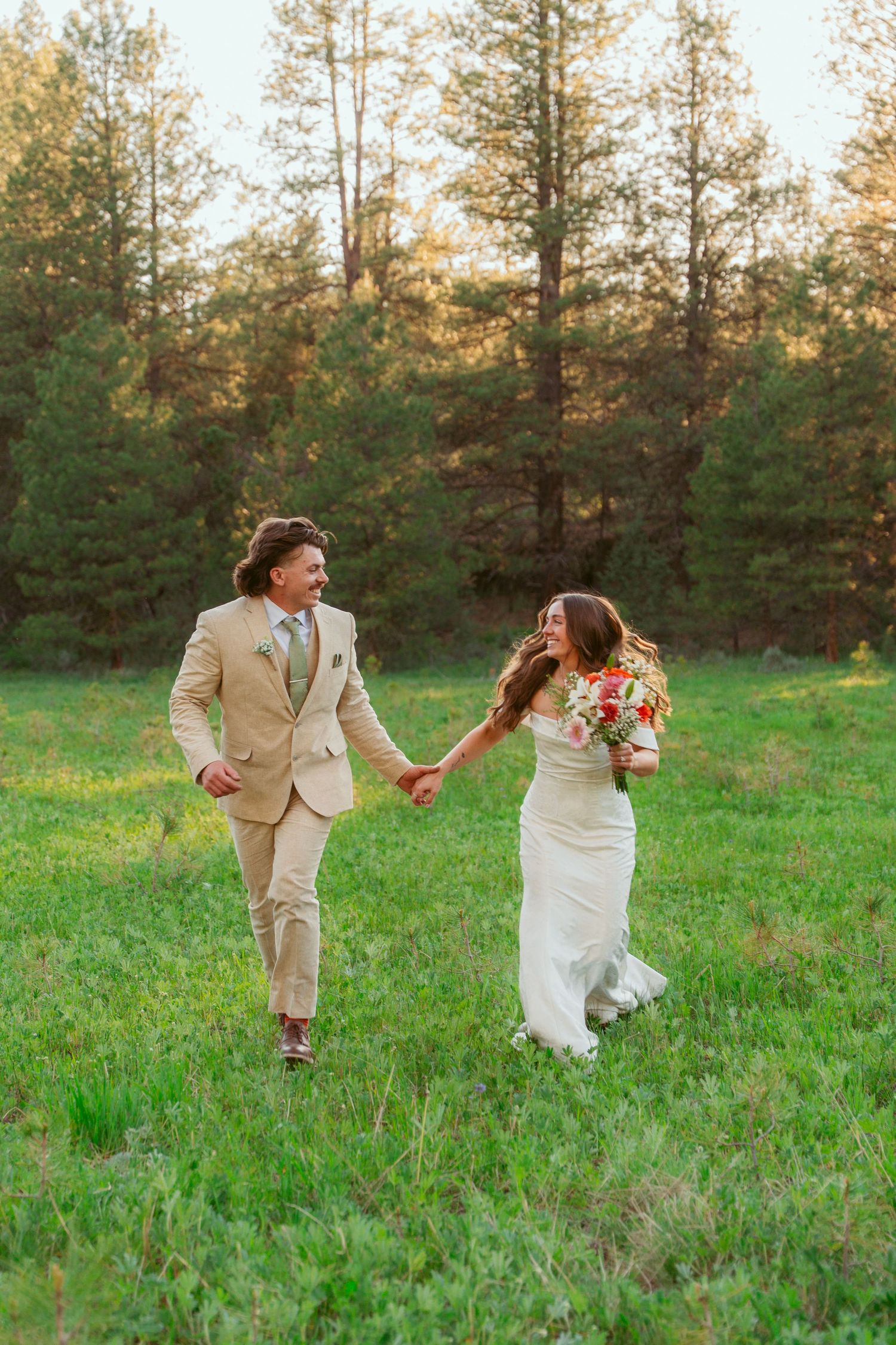 Newlyweds running playfully through a green meadow with pine trees in the background.