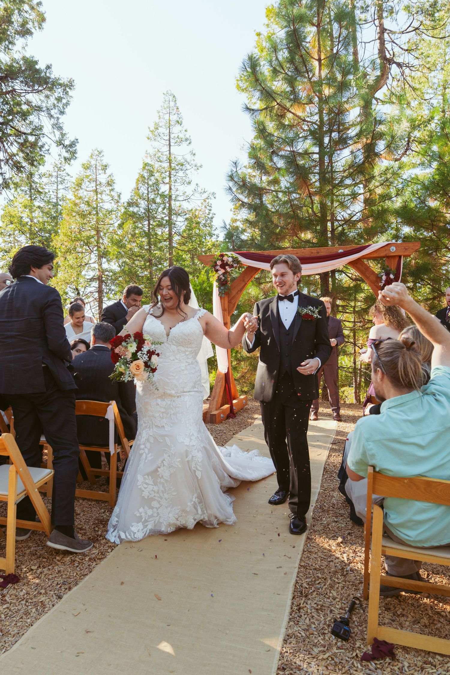 Outdoor wedding processional on a stone path surrounded by tall pine trees.