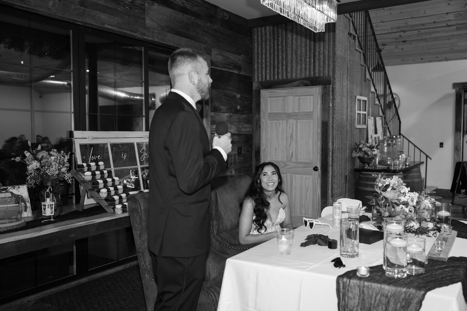 Wedding reception guests seated at decorated tables in a rustic venue with wooden beams and warm lighting.