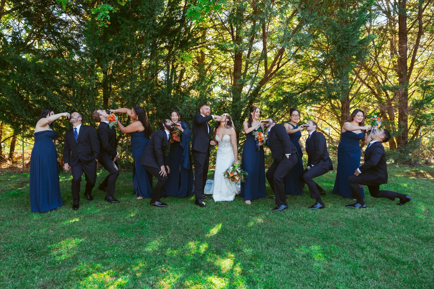 Wedding party members gather on sunny hillside for outdoor group photos in formal attire.