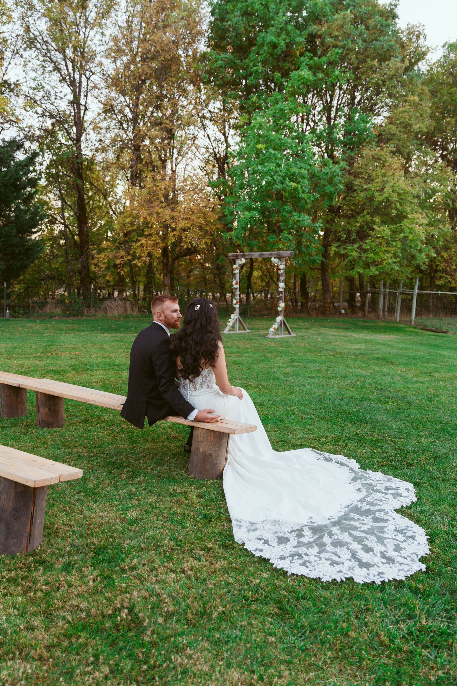 Wedding couple relaxes on wooden bench surrounded by green autumn landscape.