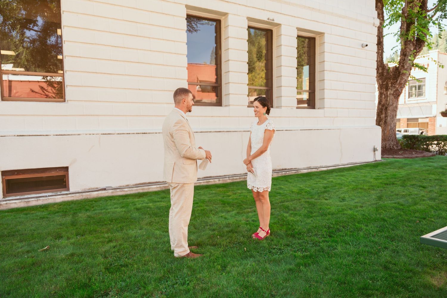 A couple stands together on a grassy lawn against a white building during their wedding day.