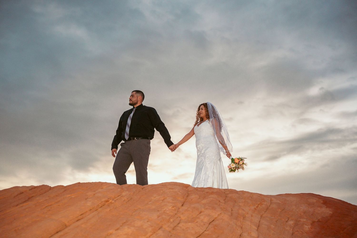 Couple walking along dramatic red rock formations during sunset in a desert landscape.
