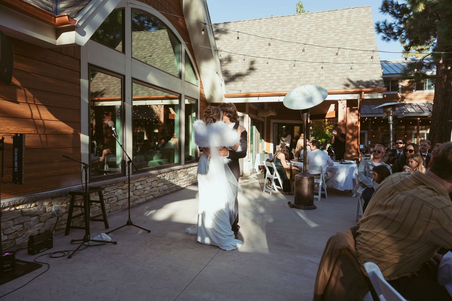 Wedding guests watch as a couple dances at an outdoor venue with rustic wooden details.