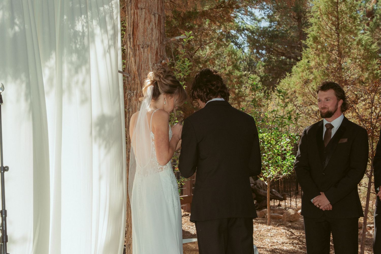 A wedding ceremony takes place outdoors with white curtains and tall trees creating a natural backdrop.