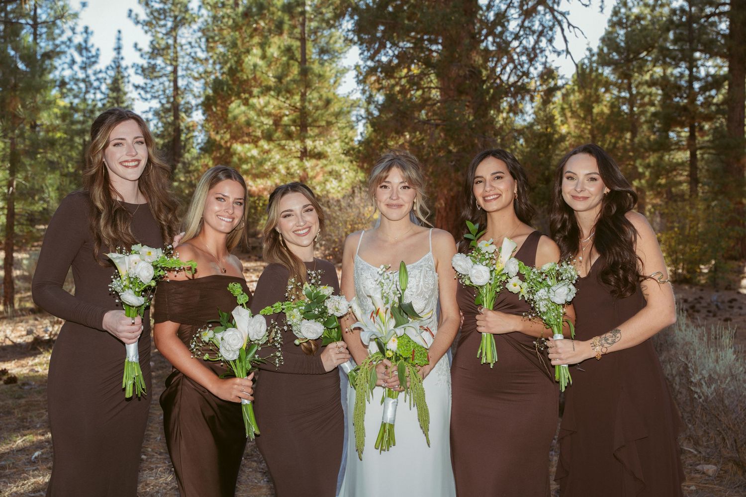 Wedding party lineup with bridesmaids in brown dresses holding white bouquets in pine forest.