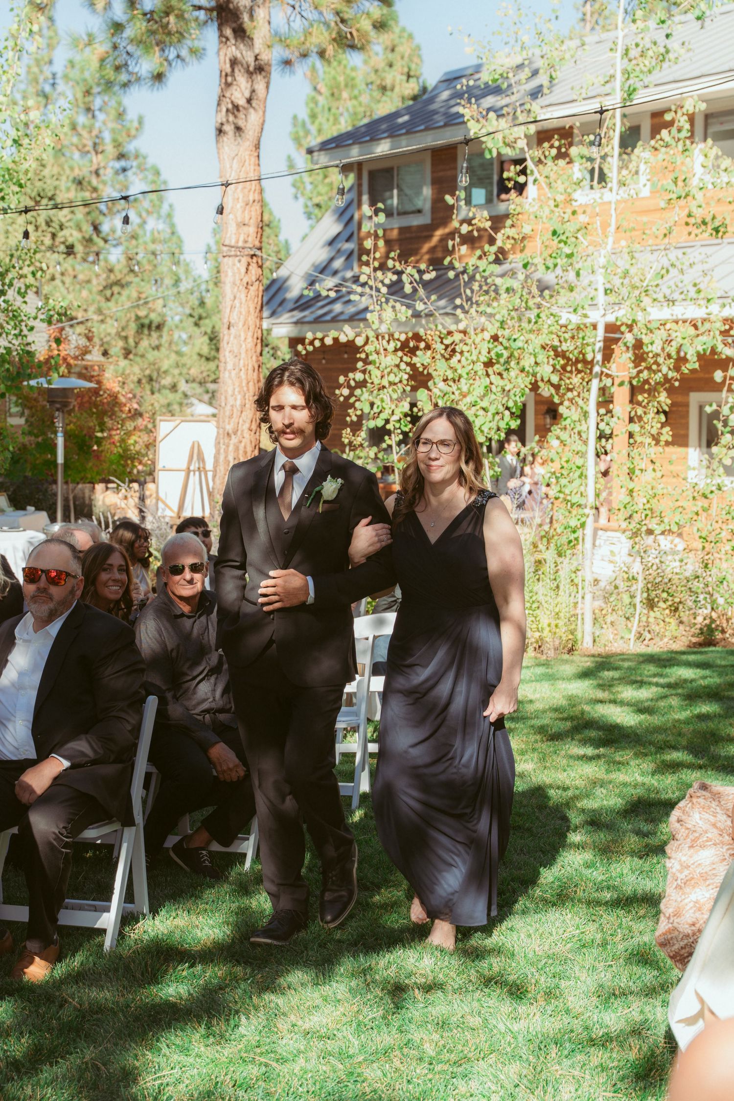 Wedding party members walk down an outdoor aisle with tall pine trees and a wooden cabin backdrop.