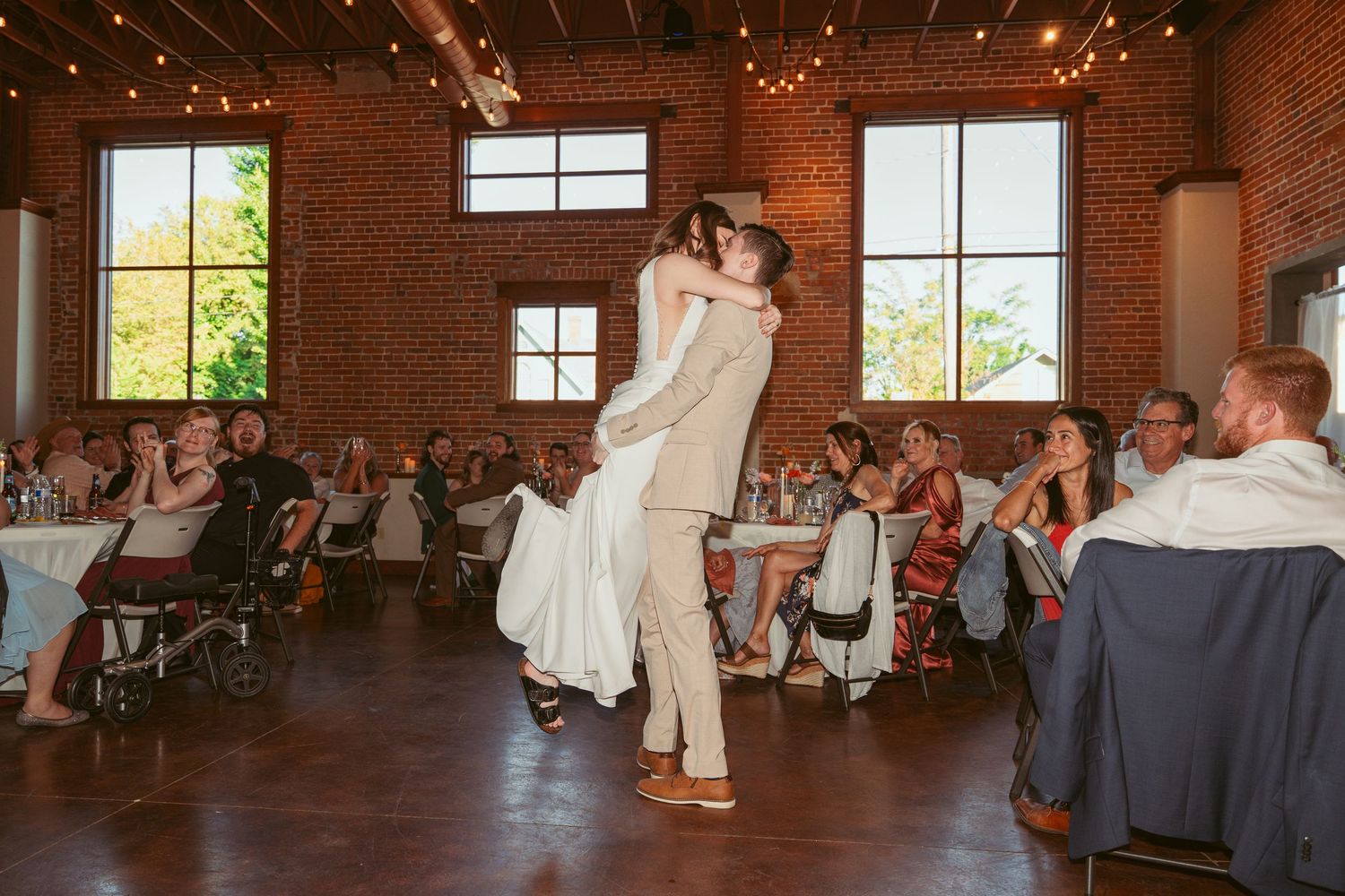 Wedding guests dance and celebrate on a rustic wooden dance floor in a brick-walled reception venue with string lights.