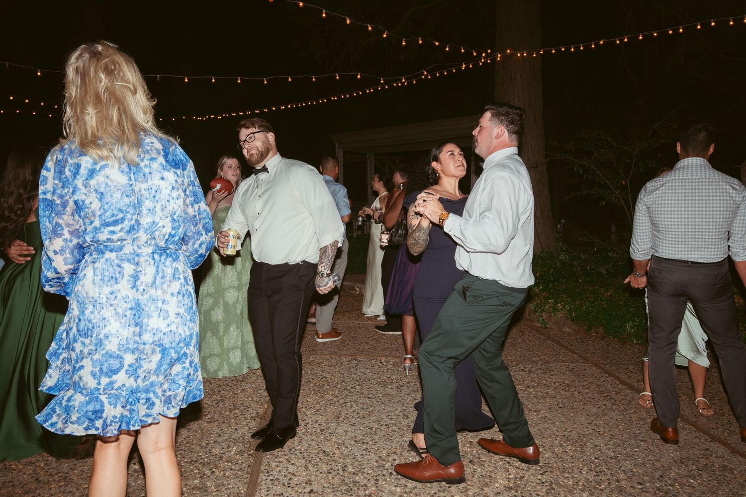 Wedding guests dancing under twinkling lights at an outdoor evening reception.