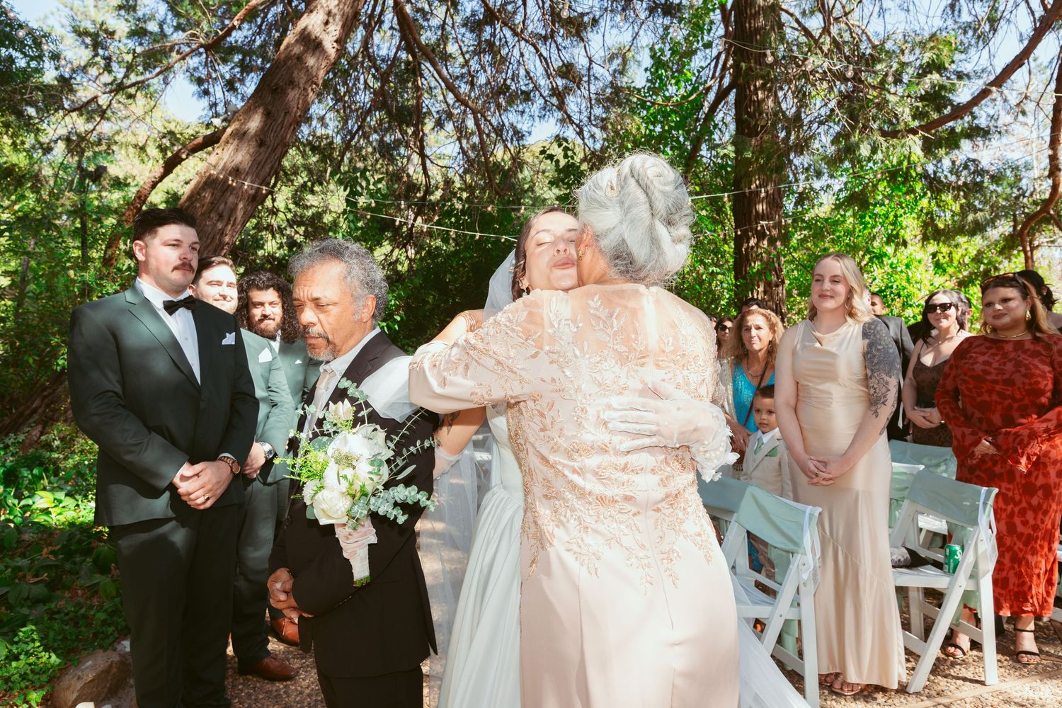 Wedding party celebrates joyfully in an outdoor garden setting with natural greenery and trees in the background.