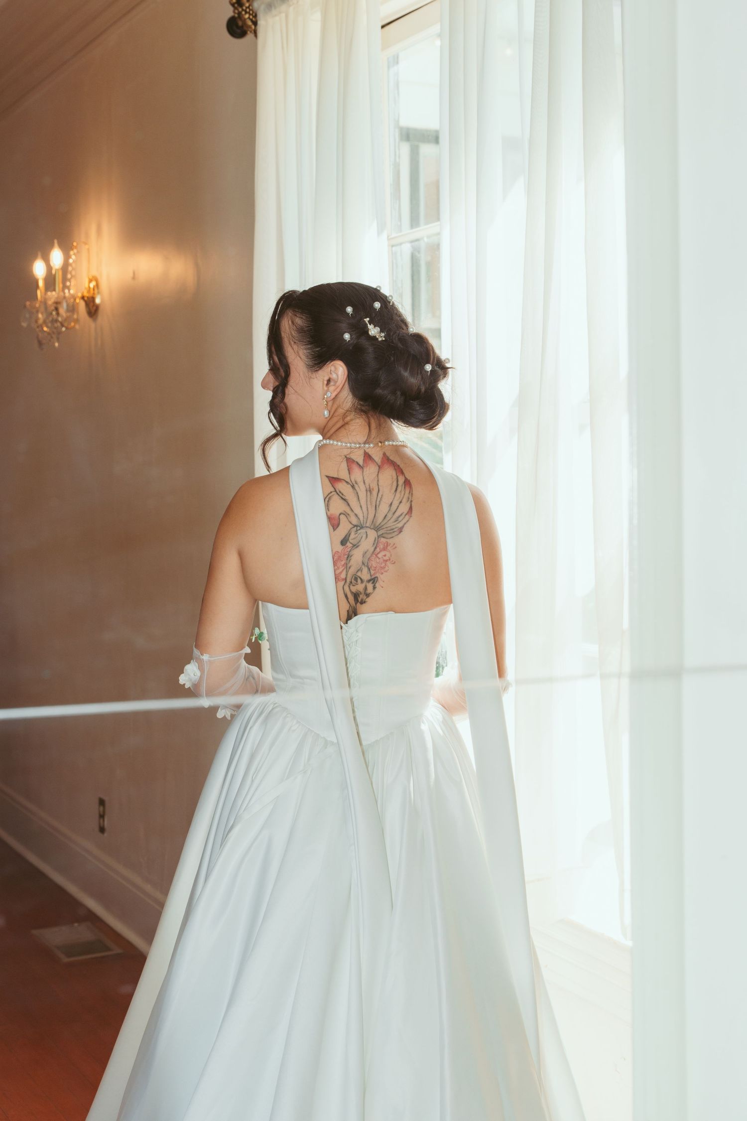 Back view of a wedding dress showing intricate tattoo details through sheer fabric near an elegant window.