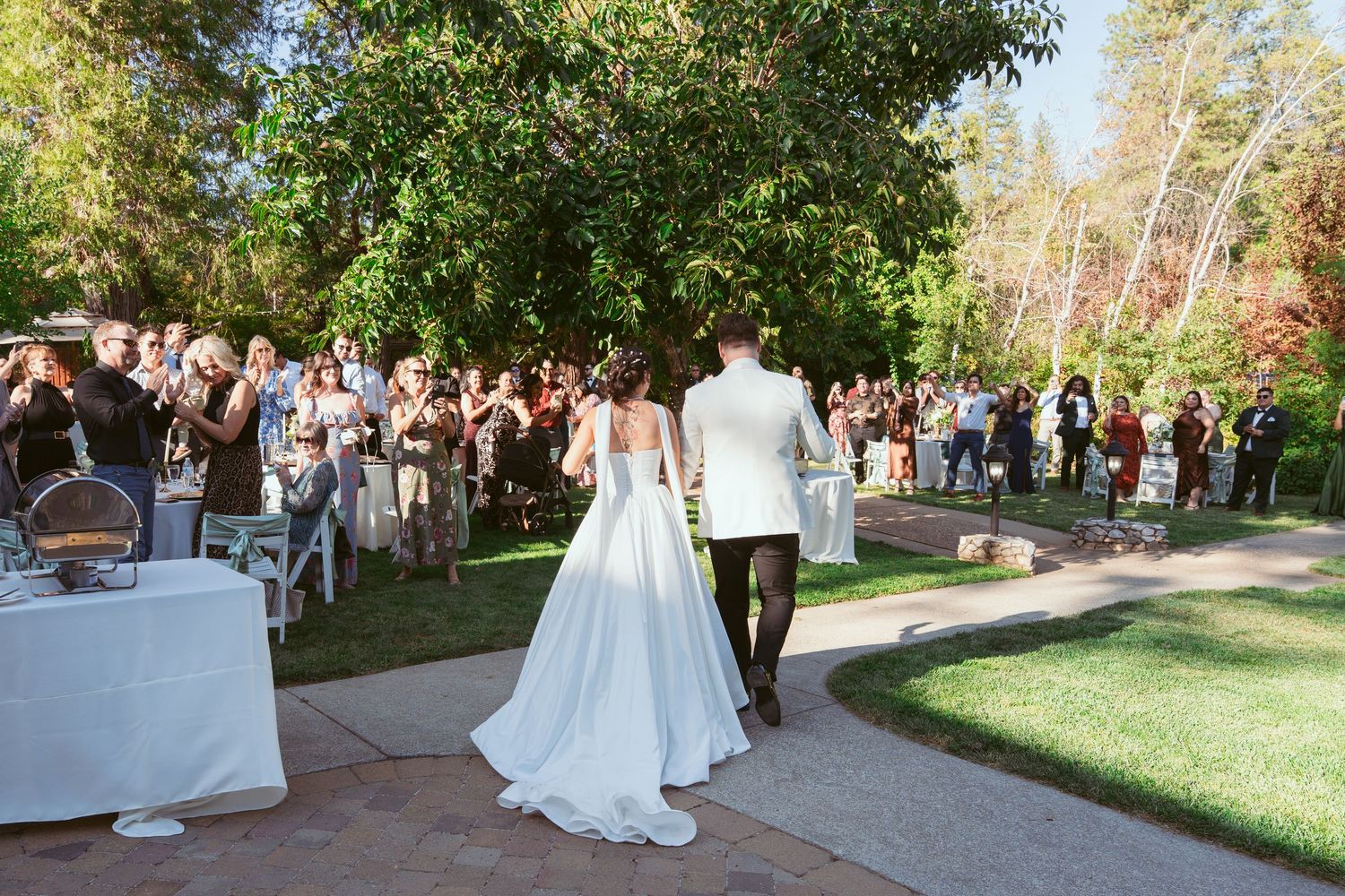 A couple in wedding attire walk along a garden path while guests gather for an outdoor reception.