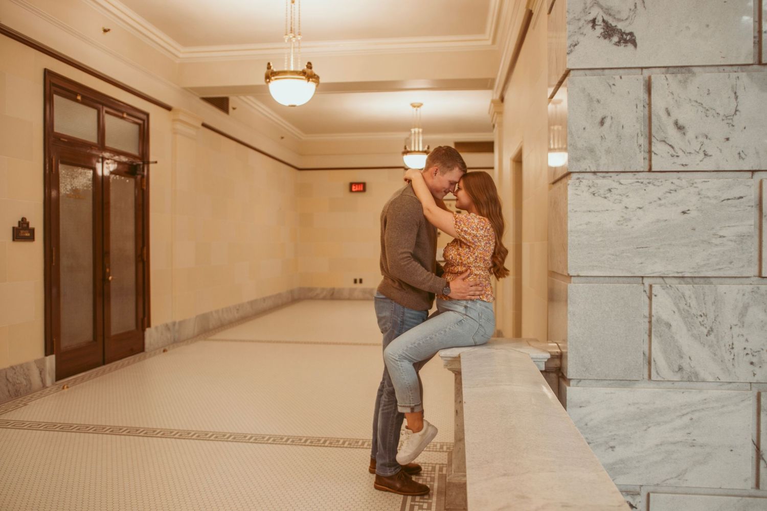 A couple shares tender moments along a marble-walled hallway in a grand building.