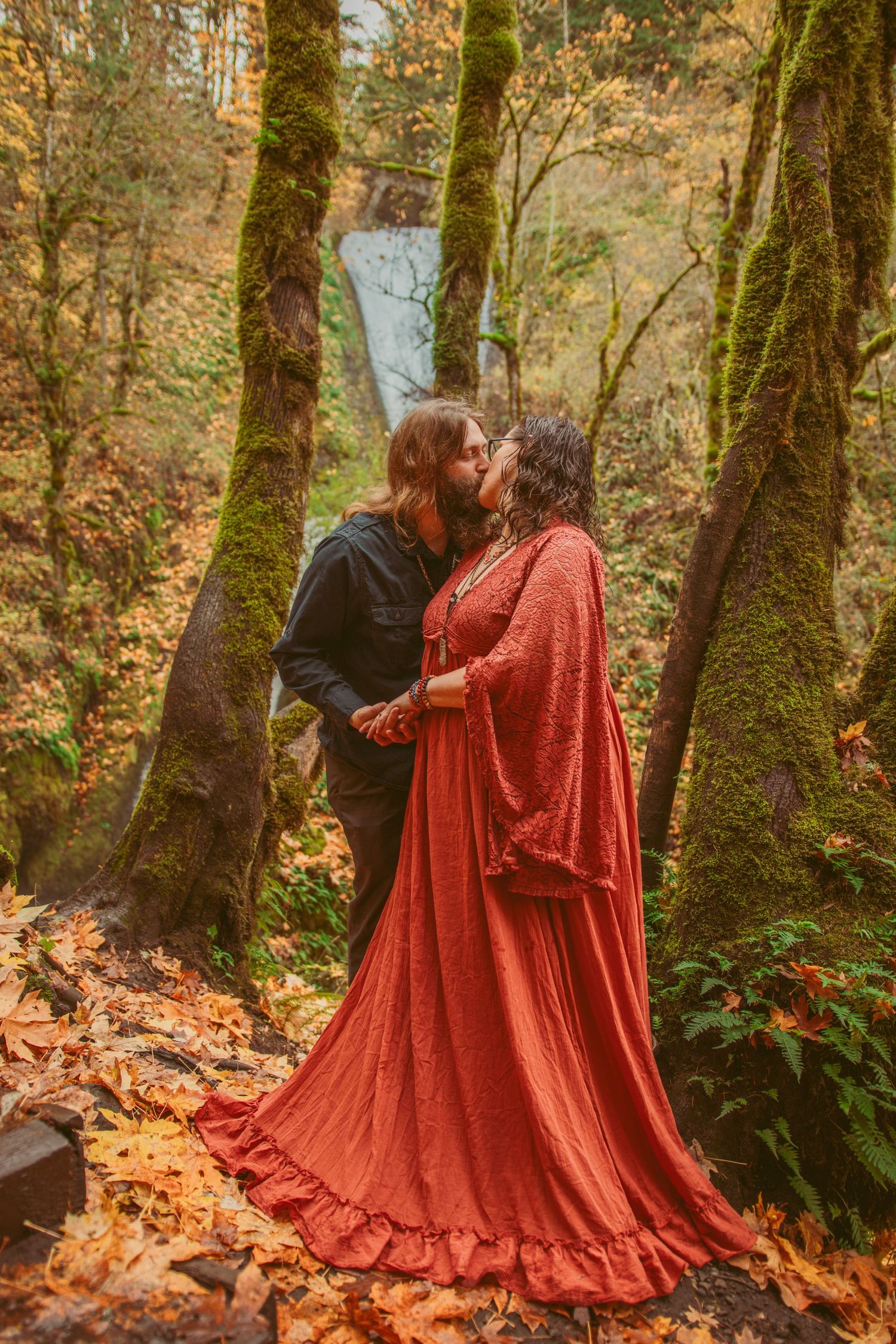 Two people share an embrace among fallen autumn leaves and mossy trees.