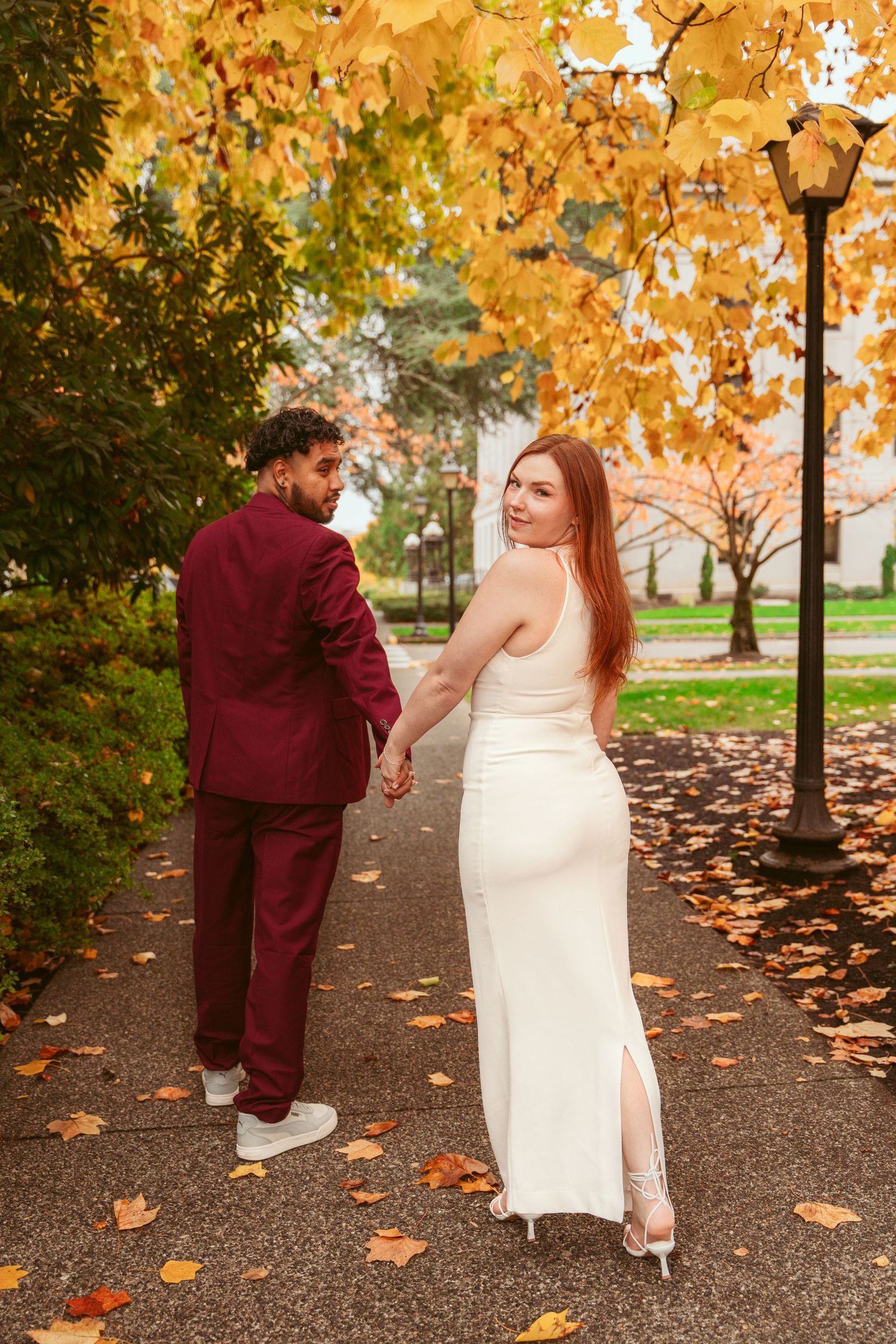 Couple in formal attire walking through a path lined with golden autumn trees.