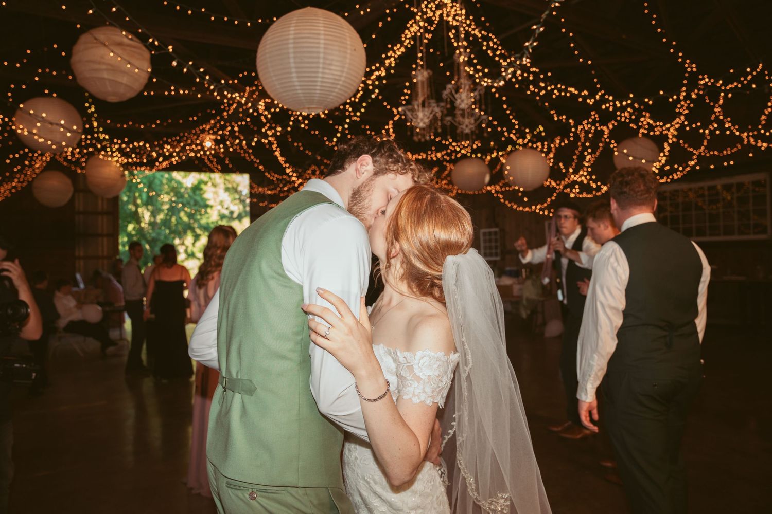 Wedding guests dancing under a canopy of string lights and paper lanterns in a rustic venue.