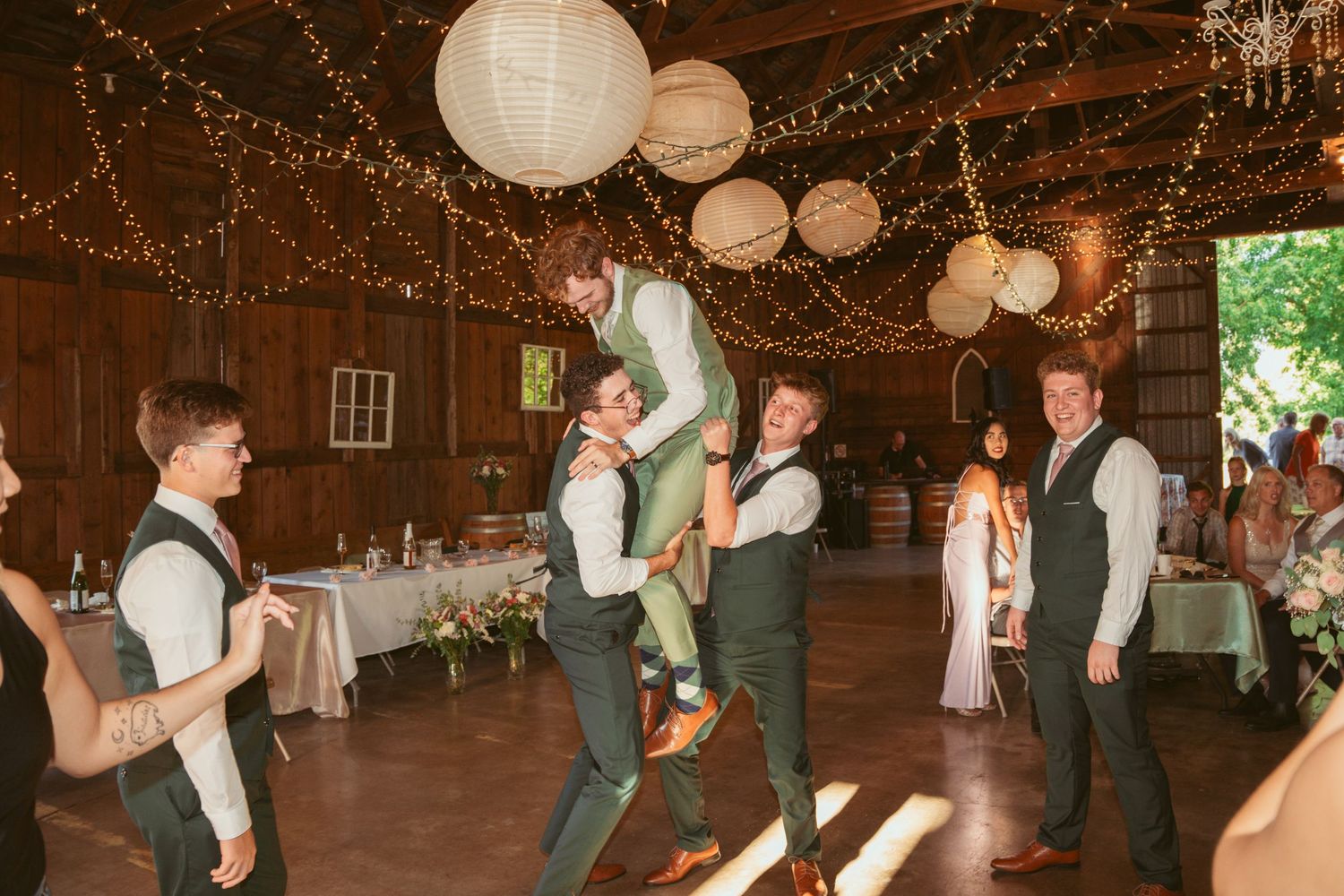 Wedding guests dance in formation during a reception in a barn decorated with string lights.