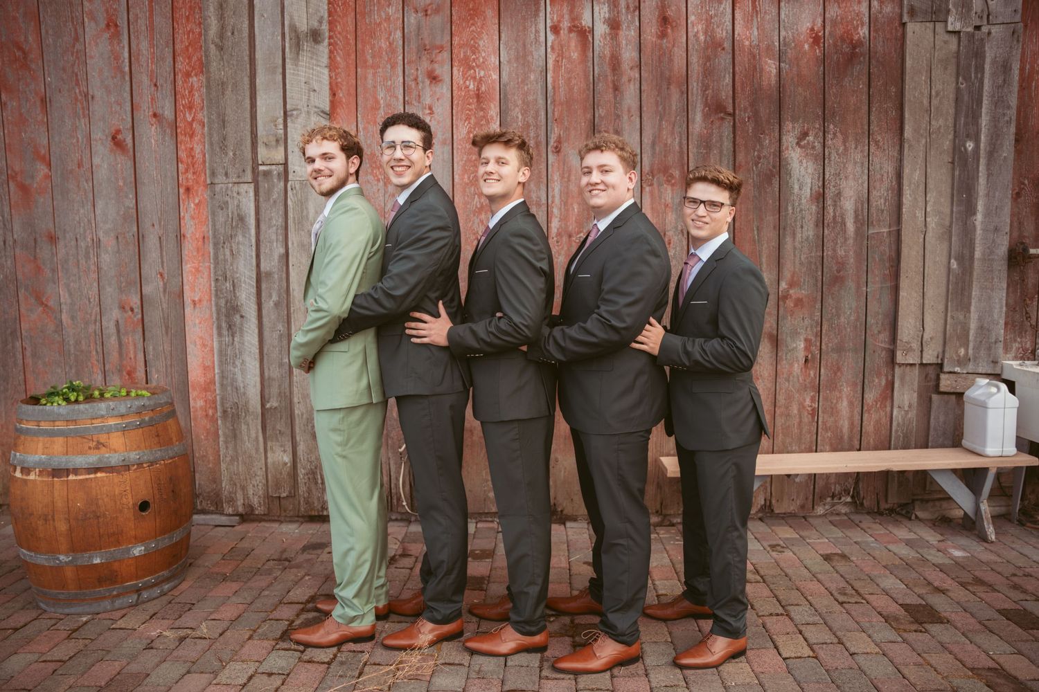 Wedding party members line up in formal suits against rustic wooden backdrop for group photos.