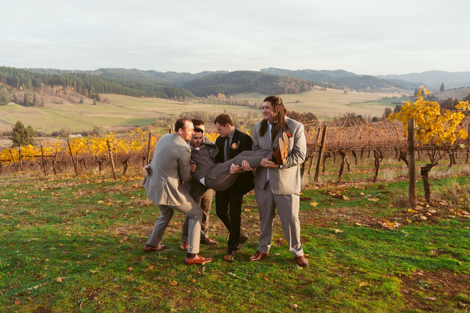 Wedding party members in grey suits playfully posing together in a vineyard setting.