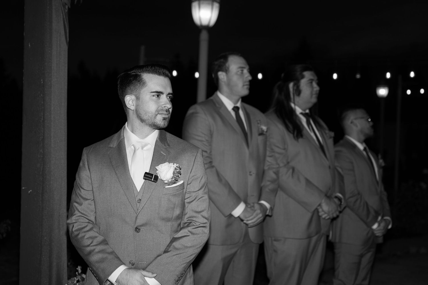 Formal wedding party in suits poses under outdoor lighting at night.