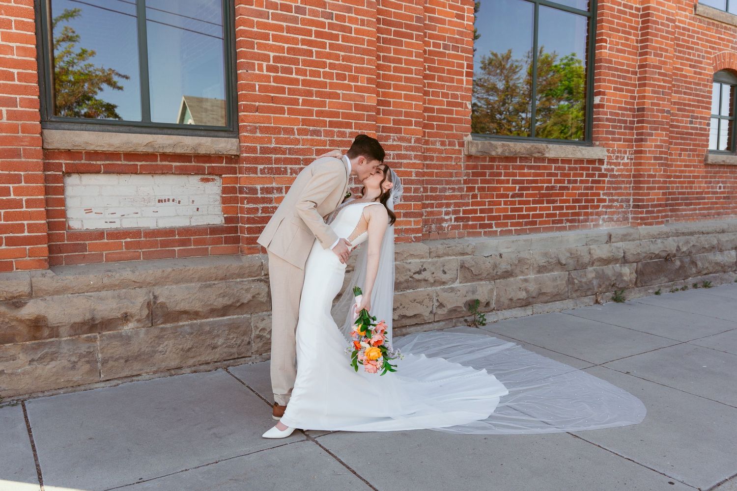 Wedding couple posing against brick wall exterior with flowing white dress and stone foundation.