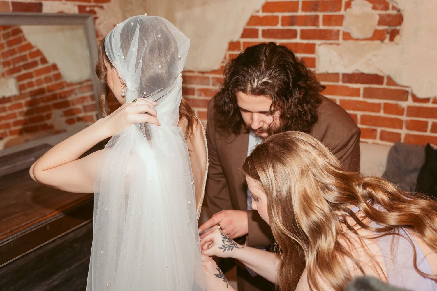Helping hands assist with adjusting a wedding veil against exposed brick wall backdrop.